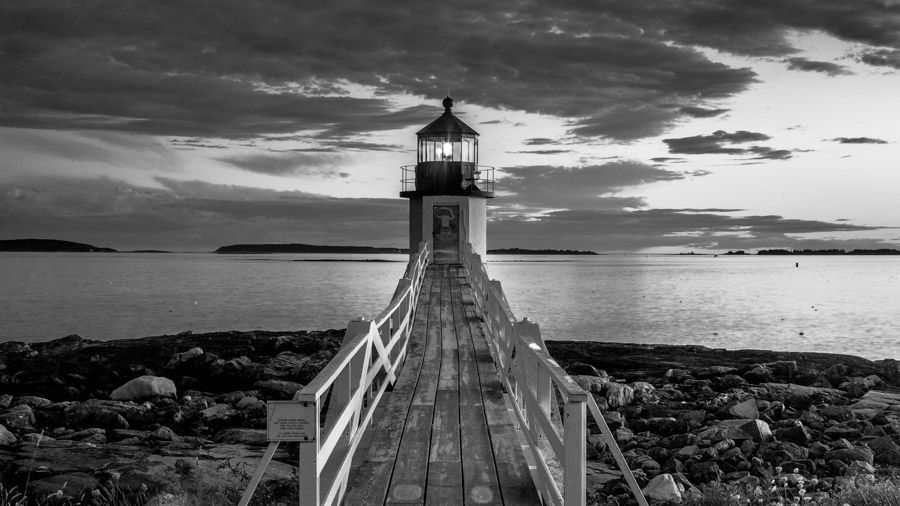 Black and white image of a lighthouse at the end of a deck overlooking the sea