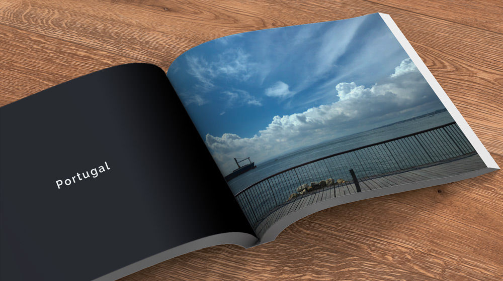 Book open on a table. Left side is a black page with text “Portugal” in white. Right side is pier, ocean, and clouds.