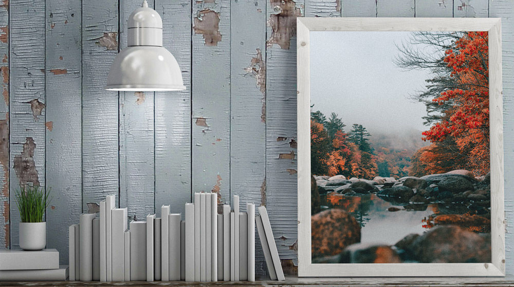 Weathered gray wall features framed print of fall nature scene with lake, large rocks, and trees with red foliage.