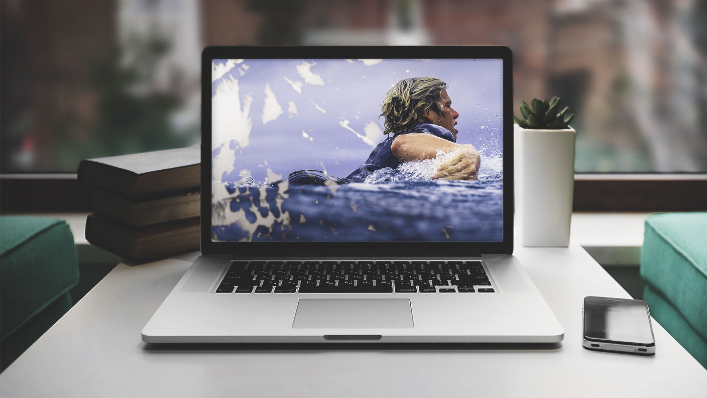 Composite image of a man swimming with palm trees in the background appears on a laptop sitting on desk next to cell phone 