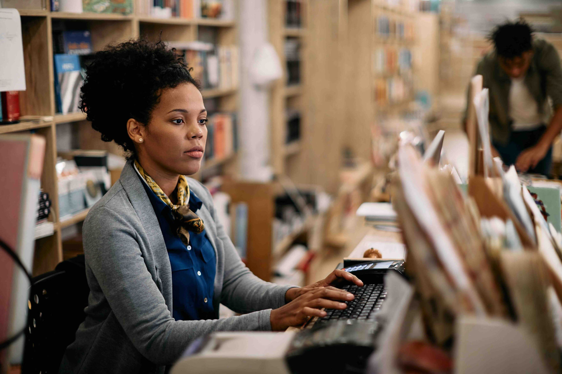 A woman in business attire typing on computer keyboard at library desk, with bookshelves in background and another person visible in distance.