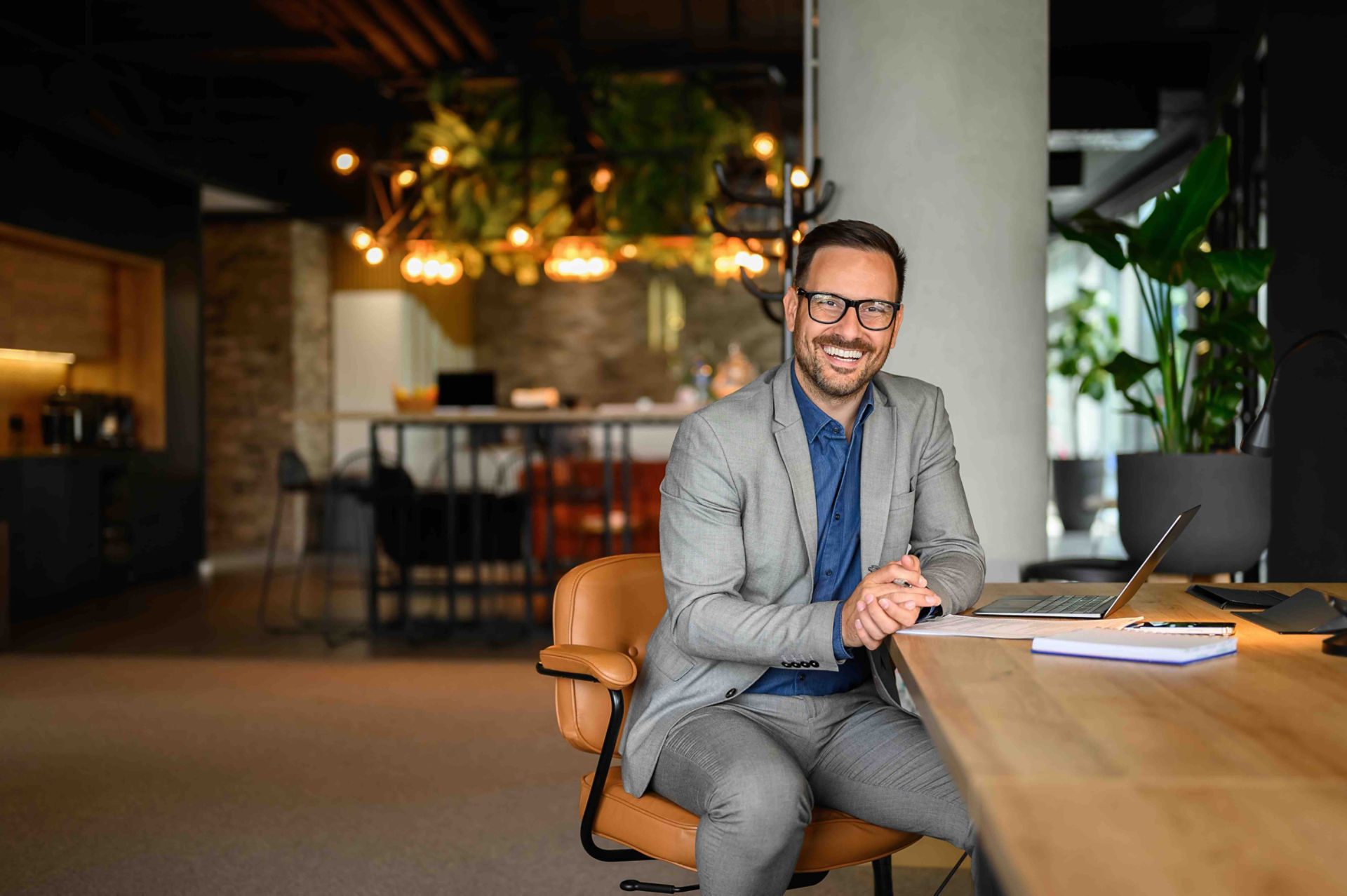 Smiling businessman in gray suit and glasses sitting at desk in stylish office with laptop, warm lighting, and plants in background.