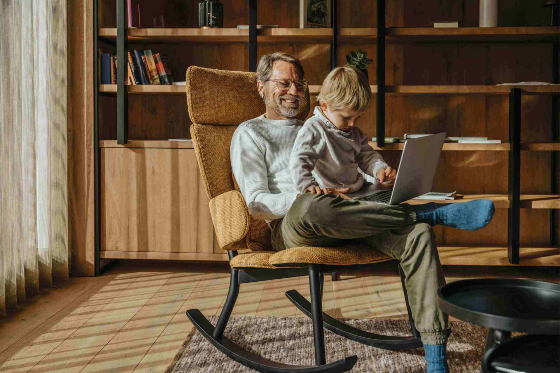 An older man and young child sit together in a rocking chair, looking at a laptop in a warmly lit room with bookshelves.