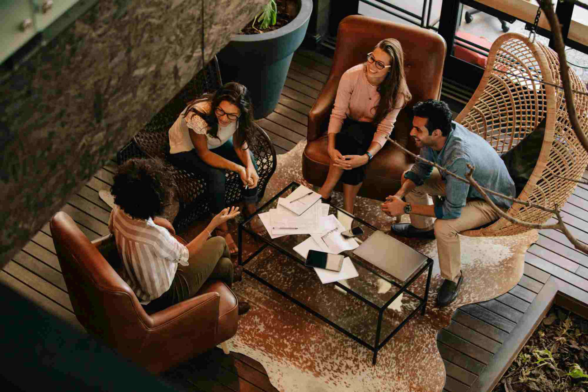 Alt text: Overhead view of four diverse professionals engaged in a casual meeting, sitting around a coffee table with papers and laptops, in a stylish office space with wooden floors and contemporary furniture.