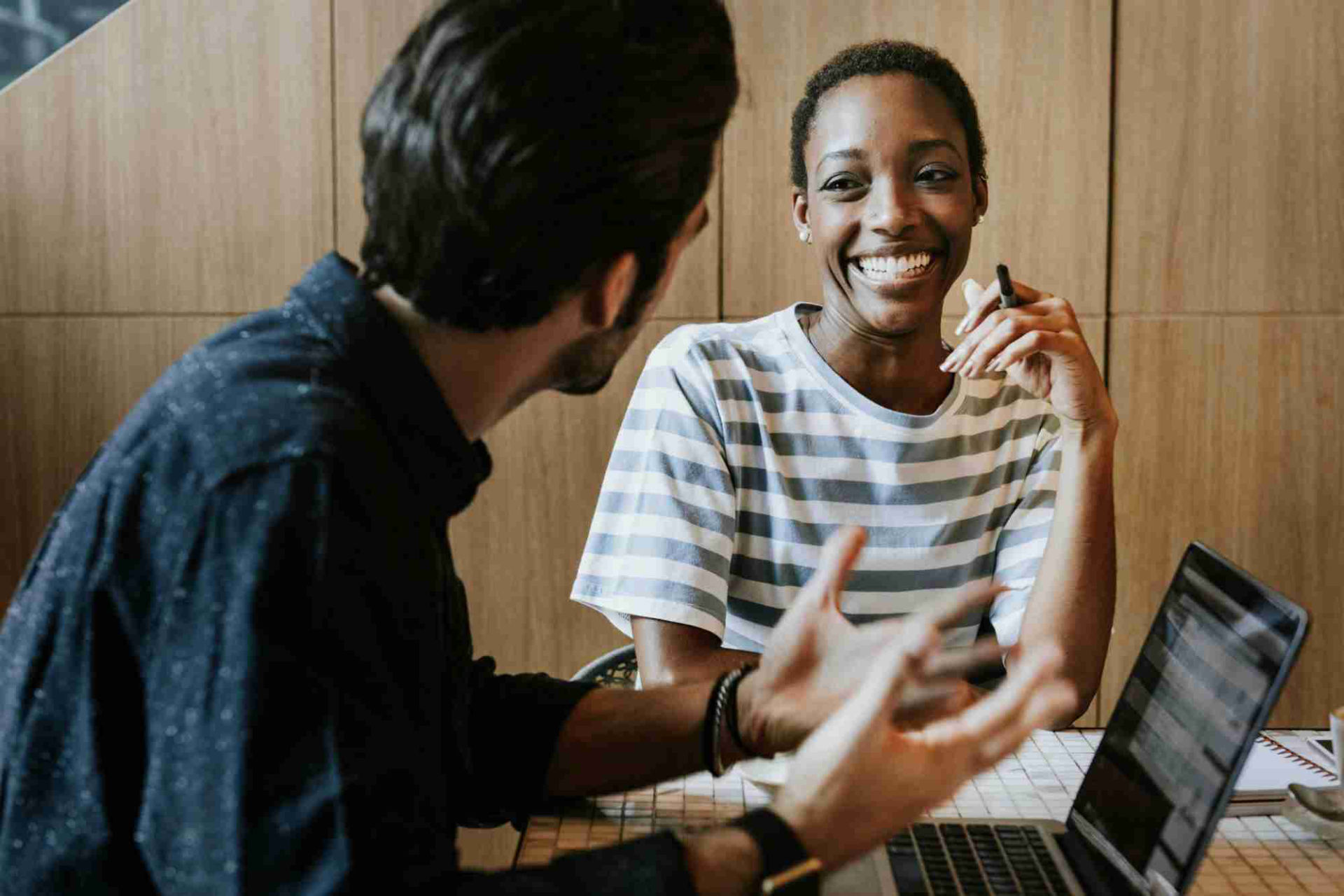 Two coworkers having an animated discussion at a desk with laptops, one person smiling broadly while the other gestures expressively, demonstrating effective workplace communication.