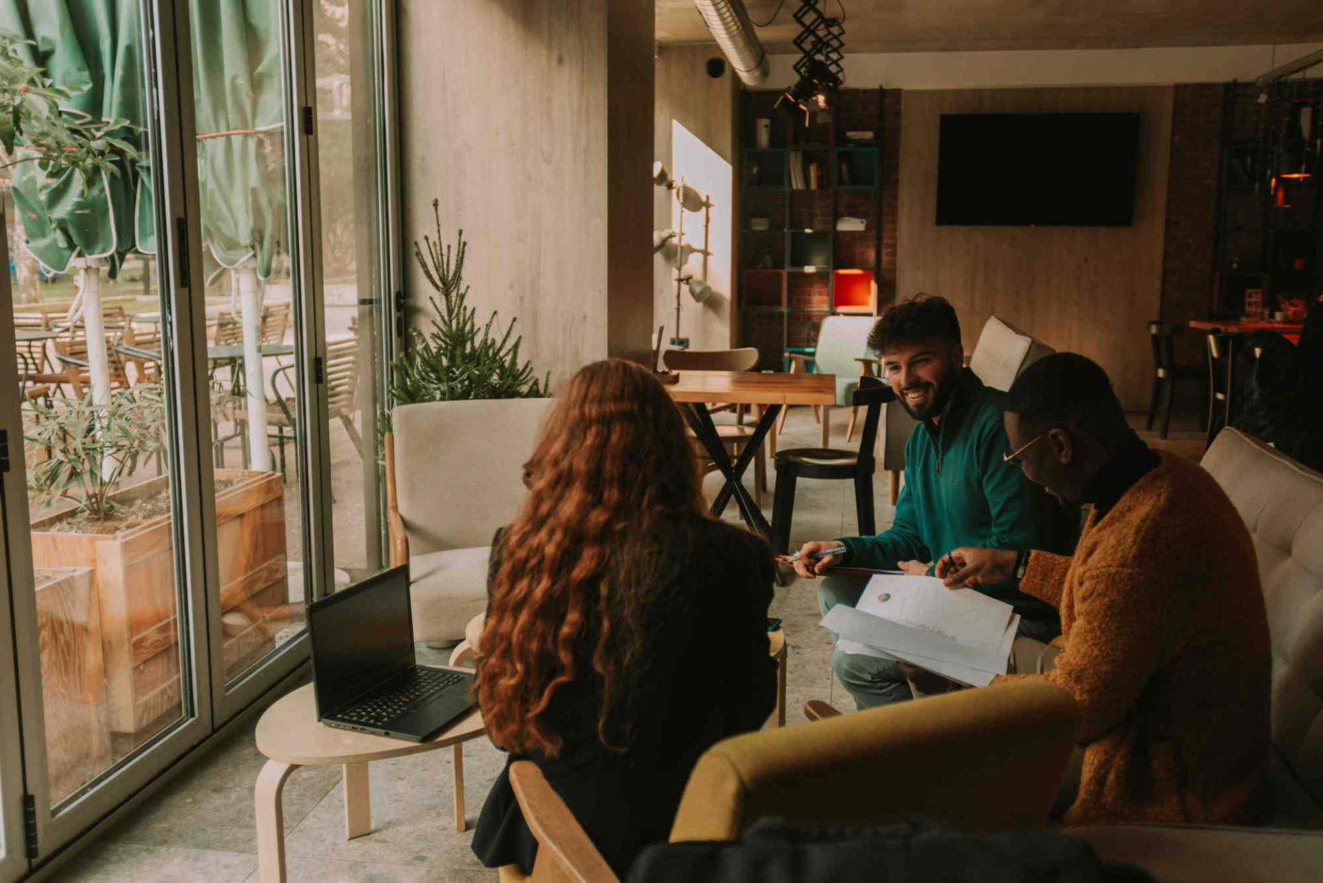 Three professionals in casual attire having a meeting in a bright office with large windows, plants, and modern furnishings. They are examining papers and engaged in discussion, showcasing teamwork and collaboration in a contemporary work environment.