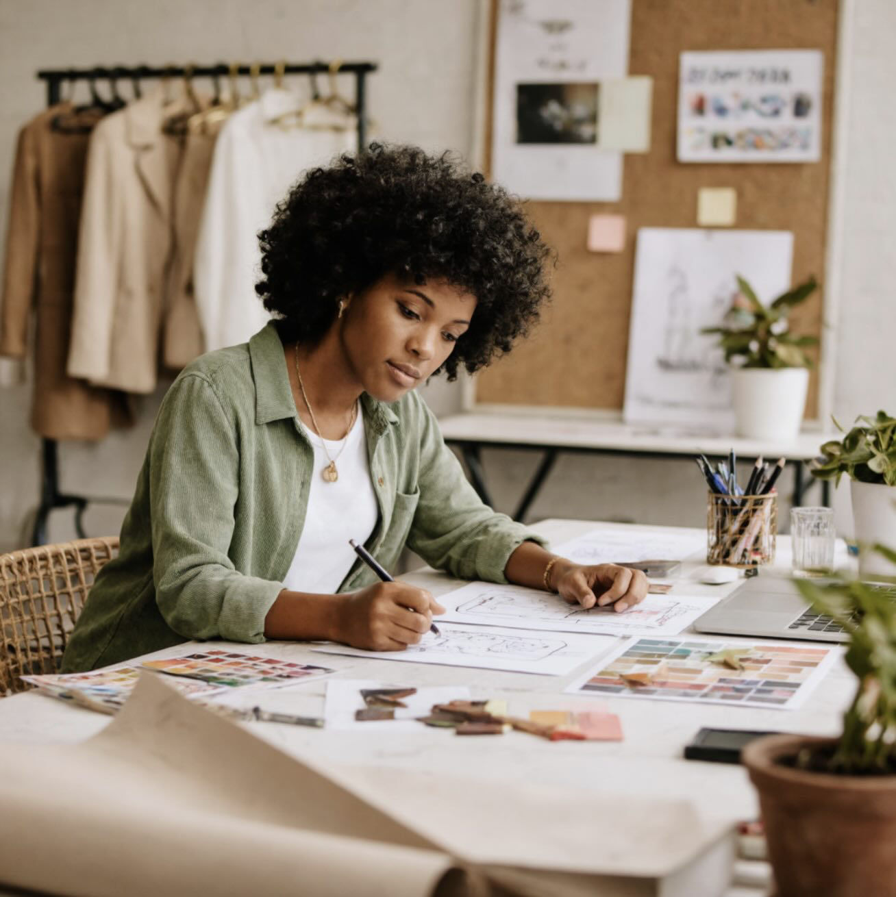 An image generated using Custom Models trained on the Firefly Image Model 4. It depicts a woman sketching designs at a desk surrounded by color swatches, mood boards, and a clothing rack, illustrating a creative workspace for fashion or interior design.