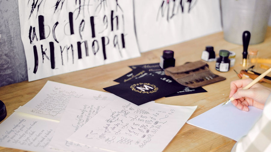 A person is practicing lettering on paper at a desk with a modern quill and ink