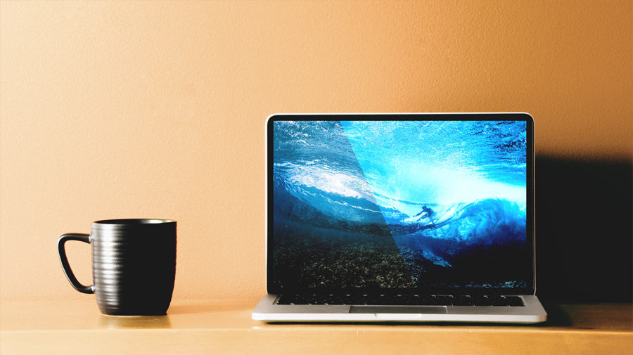 Coffee cup and laptop sitting on pale orange table. Laptop displays underwater view of surfer riding ocean wave
