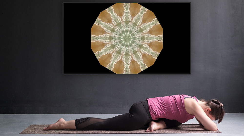 Kaleidoscope mandala appears on a monitor mounted on a gray wall. A woman practices yoga in front.