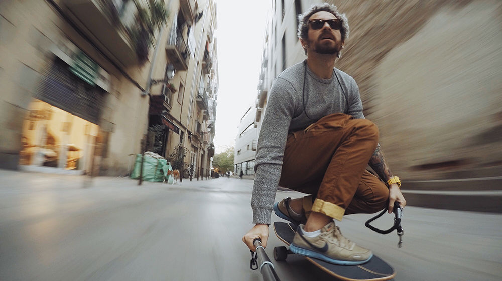 Skateboarding man with sunglasses, beard, gray sweater, and brown pants appears in front of a blurred background.