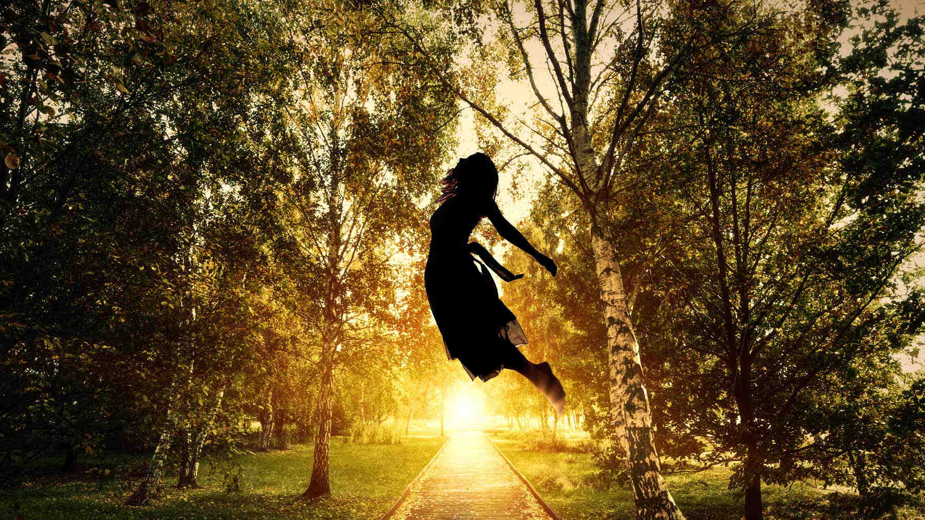 Backlit image of woman in a park jumping into the air. The sun appears below her and illuminates a walking path.