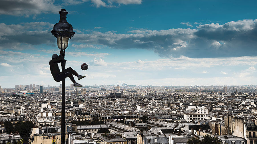 Person up on a lightpost kicking a soccer ball above a cityscape