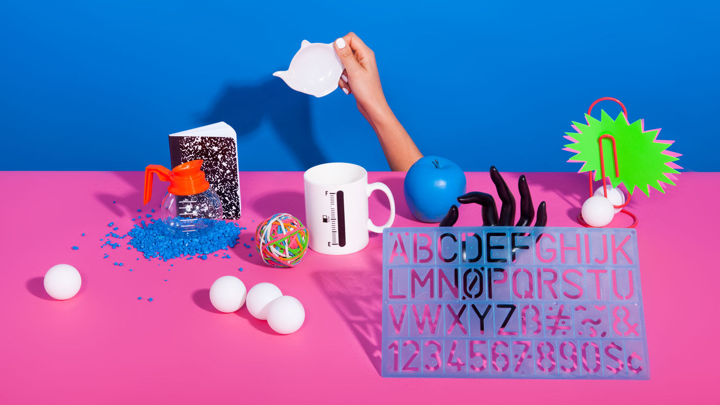 Random items such as balls, coffee pot, letter stencil, coffee cup, and plastic model hand on pink table with blue wall. 
