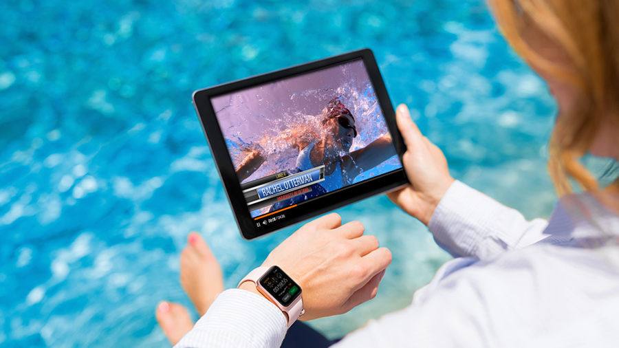 Woman at edge of swimming pool holding a tablet computer showing underwater footage of a swimmer