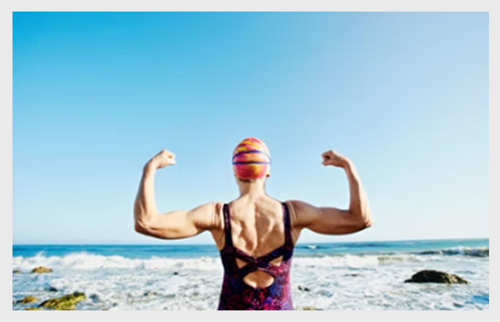 Femme senior debout sur la plage, fléchissant ses muscles avec l’océan en arrière-plan.