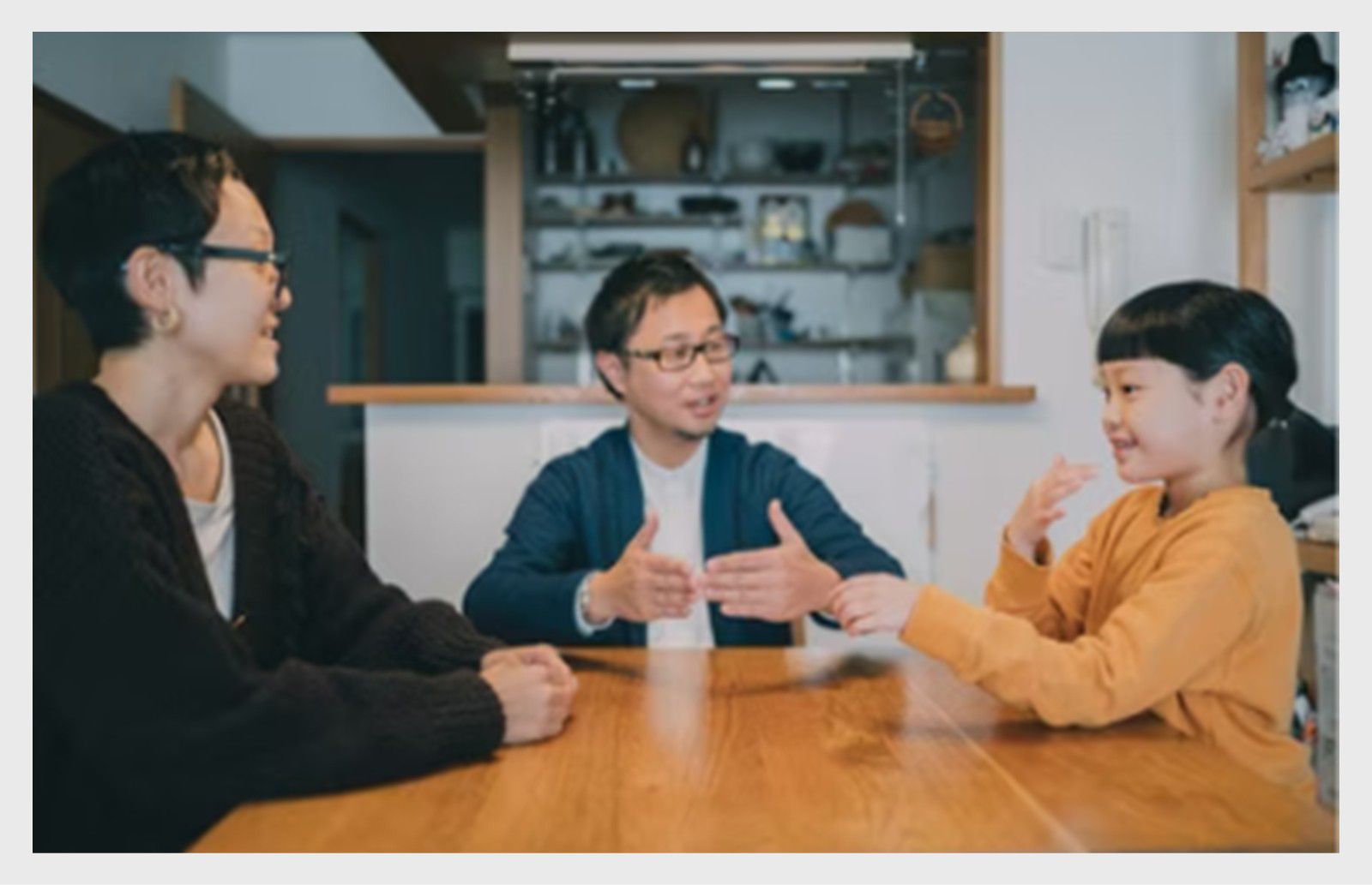 Trois personnes assises à une table à l’intérieur, engagées dans une conversation, dont une personne faisant des gestes tout en parlant.