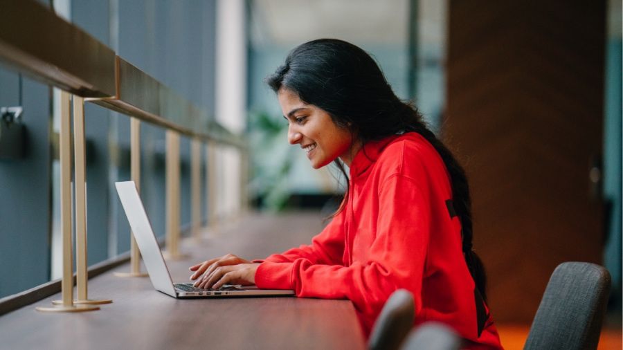 A woman with long dark hair wearing a red blouse sits at a long dark hardwood table working on a silver laptop