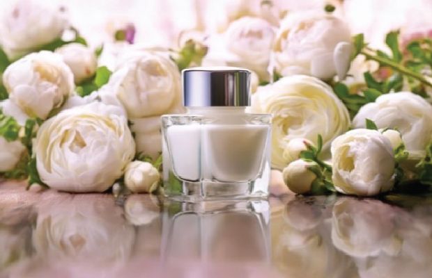 A imagem apresenta um frasco de perfume centralizado em uma superfície reflexiva, com um arranjo de rosas brancas ao fundo. Esse cenário parece ideal para um modelo de anúncio de um produto.