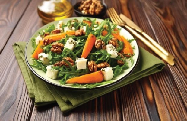 L’image représente une salade dans une assiette composée de feuilles vertes, de légumes, de morceaux de fromage blanc et de noix. Des condiments comme de l’huile d’olive et des noix se trouvent en arrière-plan. Des couverts dorés sont posés à côté de l’assiette. Cette image pourrait servir d’élément visuel pour des créations publicitaires.